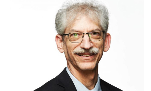 Portrait of a person with short gray hair and glasses, wearing a dark suit jacket over a light-colored shirt, set against a plain white background.