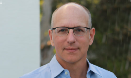Portrait of a person with short hair wearing glasses and a light blue button-up shirt, standing outdoors with a blurred background featuring greenery.