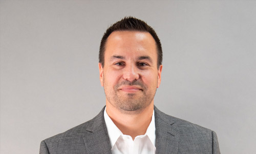 Portrait of a person with short dark hair wearing a grey suit jacket over a white dress shirt, set against a plain light grey background.