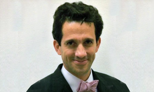 Portrait of a person with short dark hair wearing a formal black suit, white dress shirt, and light-colored bow tie, set against a plain light background.