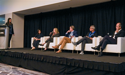 Five panelists seated on stage in white chairs during a professional conference, with a moderator standing at a podium to the left and a black curtain backdrop.