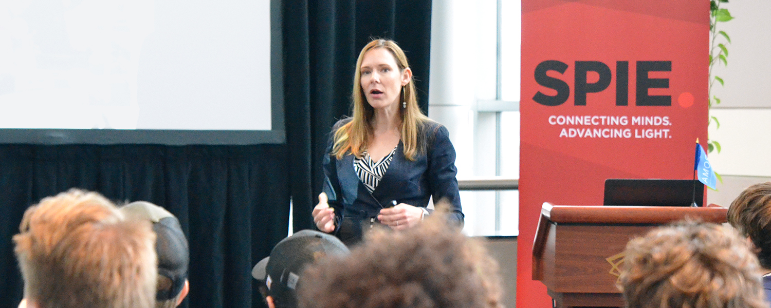 A person presents at the front of a conference room, with a lectern and microphone to the right and a large red vertical banner reading “SPIE – Connecting Minds. Advancing Light.” in the background. Attendees are seated in the foreground.