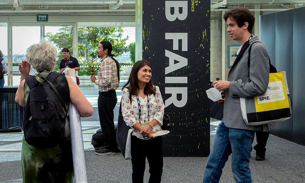 In the foreground, three individuals are conversing—one with a backpack and rolled-up poster, another smiling and listening, and a third person holding papers and a bag labeled "SPIE." A vertical sign reading "JOB FAIR" is visible with other attendees in the background.