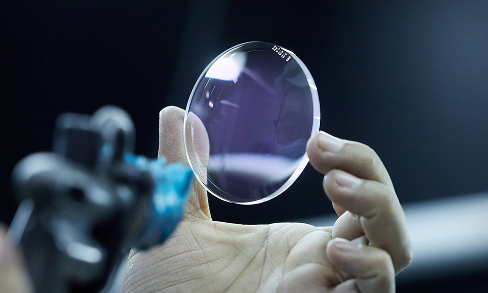 Hand holding a clear, round optical lens with a slight purple tint, positioned near a spray tool against a dark background. The lens reflects light, highlighting its smooth surface and precision edges.