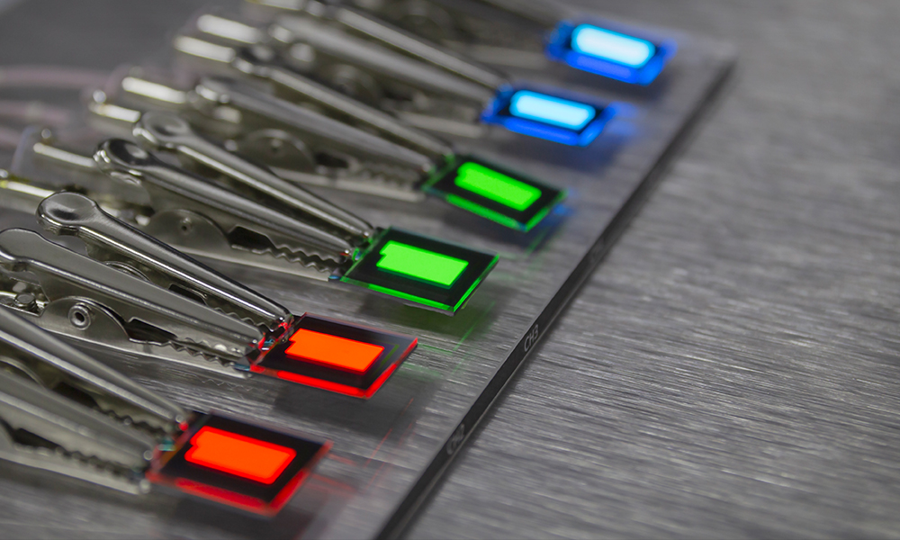 Close-up view of several small rectangular light panels in red, green, and blue, each held by metallic alligator clips attached to a flat surface. The panels are evenly spaced in rows, and the background is a brushed metal texture.