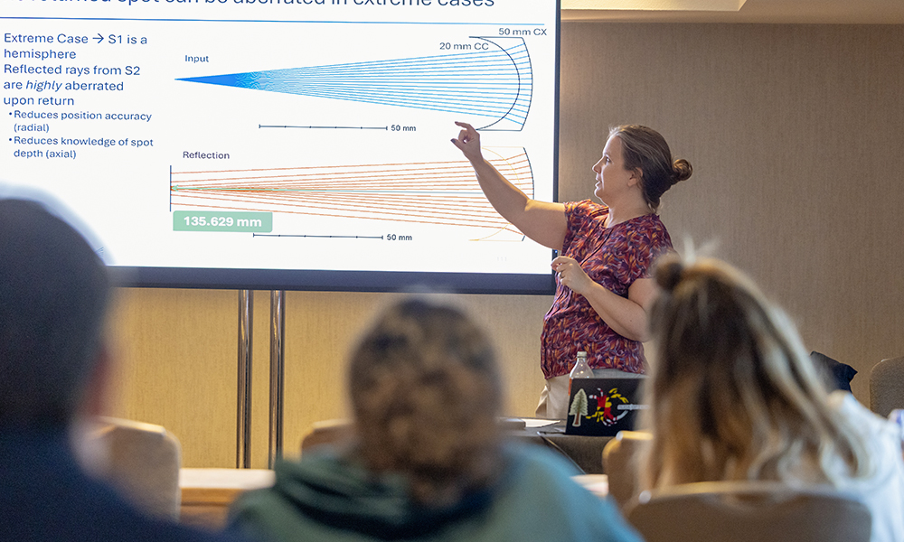 Person giving a presentation in a conference room, pointing at a slide on a large screen that shows diagrams and text about optical reflections and measurements. Several attendees are seated and watching the presentation.