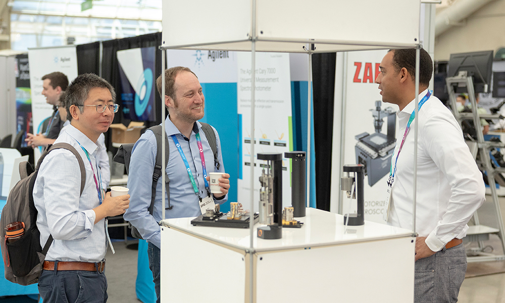 Three conference attendees stand and talk at a technology exhibition booth, with scientific instruments displayed on the counter and company banners visible in the background at SPIE Optics + Photonics exhibition