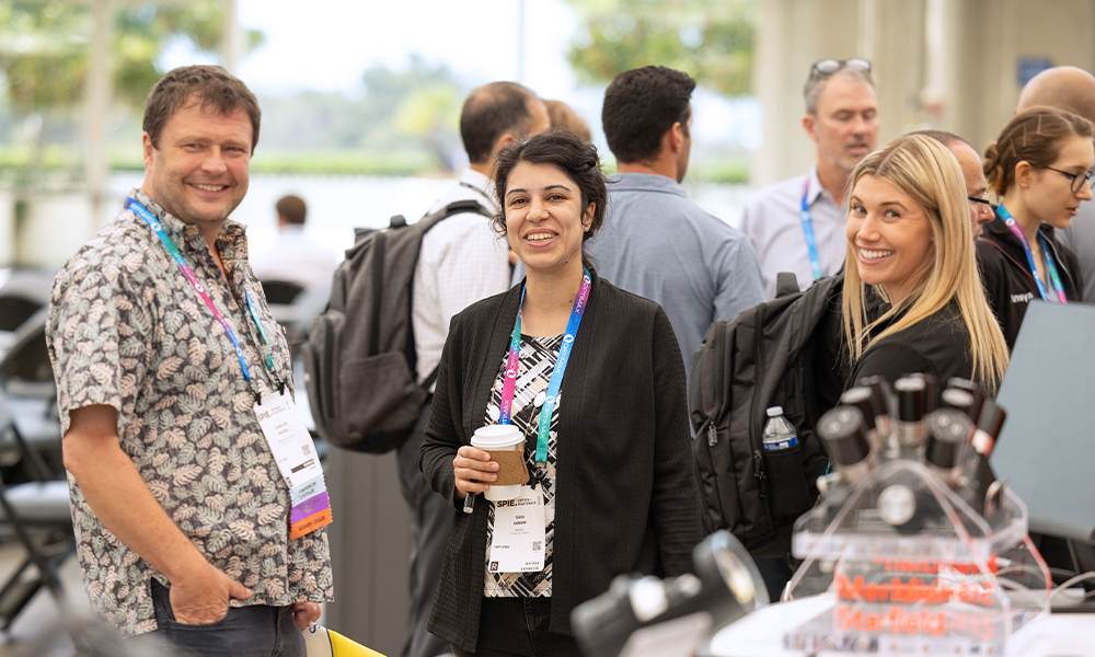 Group of people standing and conversing at Optics and Photonics, wearing name badges and lanyards, with one person holding a coffee cup. 