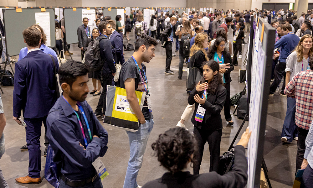 Poster session in a large conference hall with attendees standing and discussing research displayed on boards. Multiple rows of posters are visible, and people are engaged in conversations throughout the space.