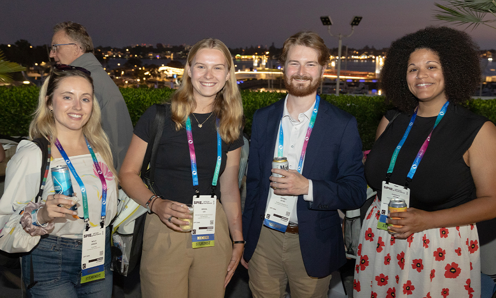 roup of four SPIE Optics + Photonics attendees wearing event badges and smiling at an outdoor evening reception, with a cityscape in the background and one additional person visible behind them.