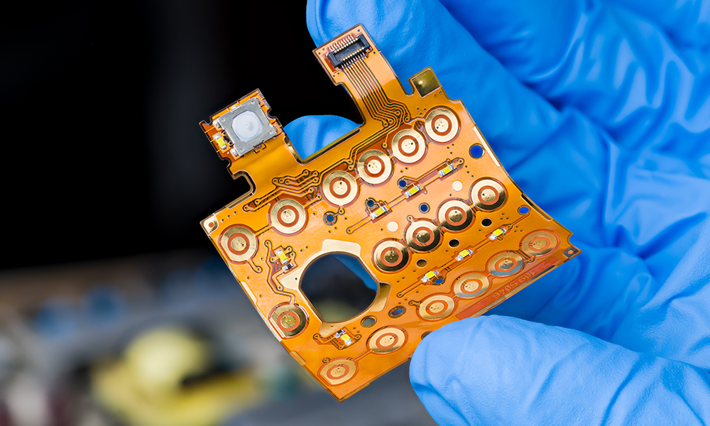 A gloved hand holding a flexible orange circuit board with multiple electronic components.