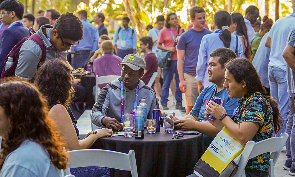 Outdoor networking event with multiple people seated at round tables and others standing in groups. Tables have drinks and bottles, and the setting includes trees and natural light, suggesting an open-air venue.