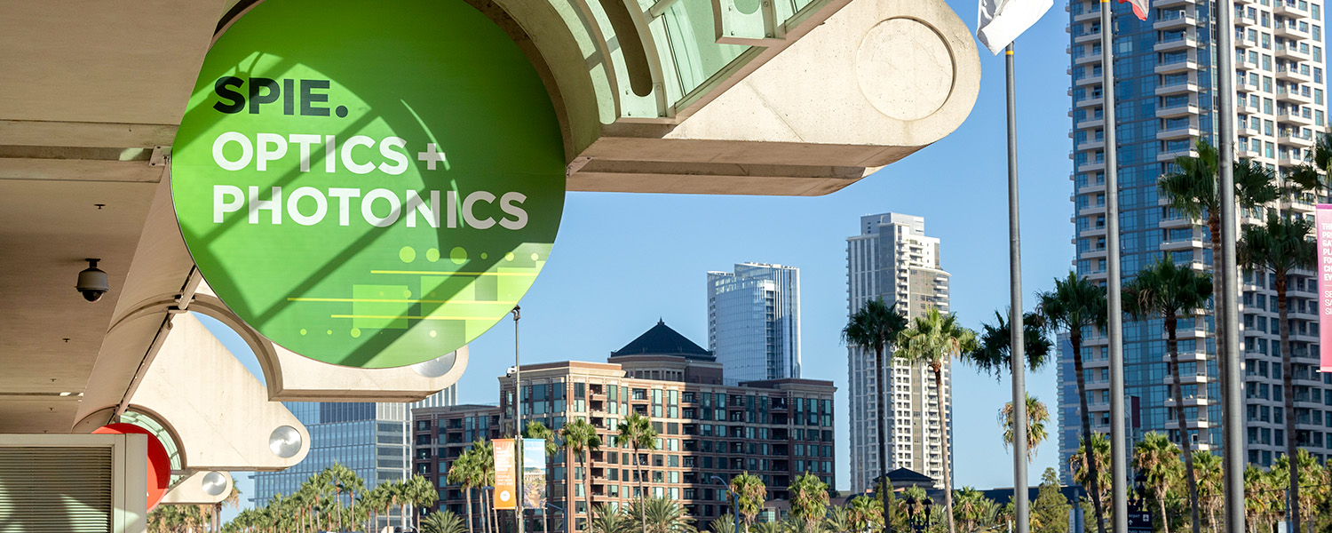 Large green circular sign reading “SPIE. OPTICS + PHOTONICS” mounted on the exterior of a building, with palm trees and tall modern skyscrapers in the background under a clear blue sky. 