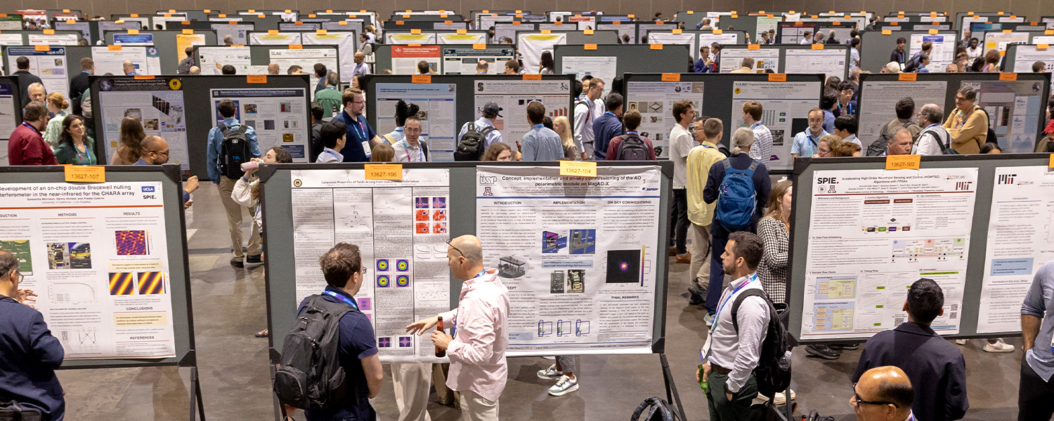 Large indoor poster session with rows of research posters displayed on boards, each featuring text, charts, and images. Numerous attendees are standing and walking through the aisles, some reading posters and others engaging in discussions.