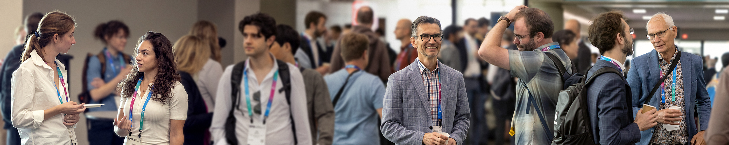 People networking in a bright conference hallway, wearing name badges and talking in small groups during SPIE Optics + Photonics,