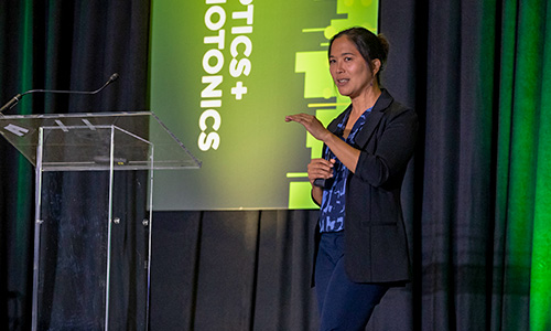  Person giving a presentation on stage next to a clear podium, with a green and black backdrop displaying the SPIE Optics + Photonics Logo.