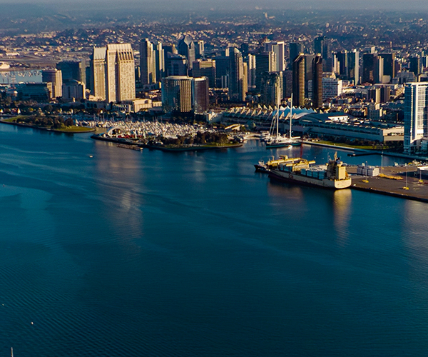 Aerial view of  the city of San Diego with a dense cluster of skyscrapers along the waterfront, an active harbor with docks and ships, and a bridge spanning part of the bay, with mountains faintly visible in the distance.