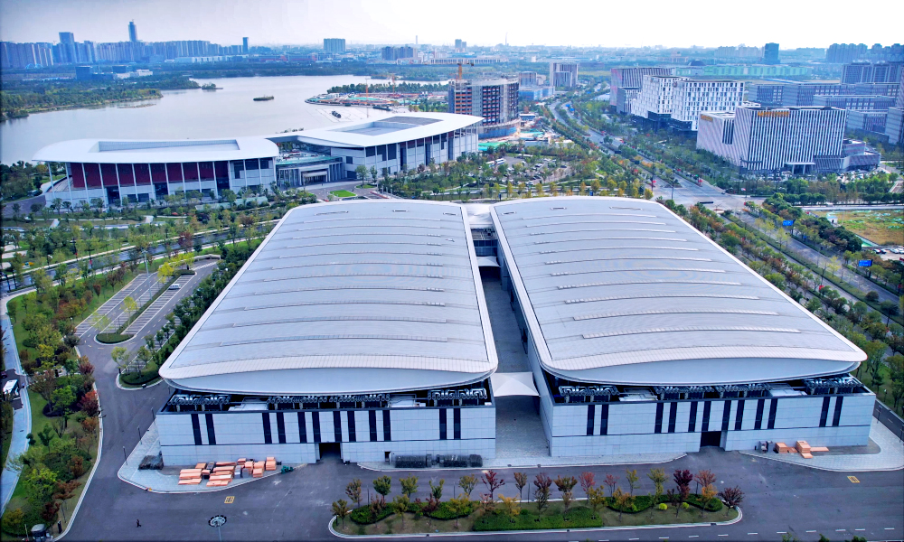 Aerial view of Nantong International Convention Center with two large, curved-roof buildings, surrounded by landscaped roads, nearby waterfront, and high-rise buildings in the distance.
