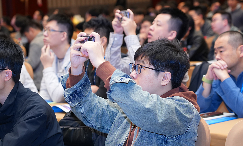 An audience seated in a large conference hall, with attendees listening attentively and several people raising smartphones to take photos or record the presentation.
