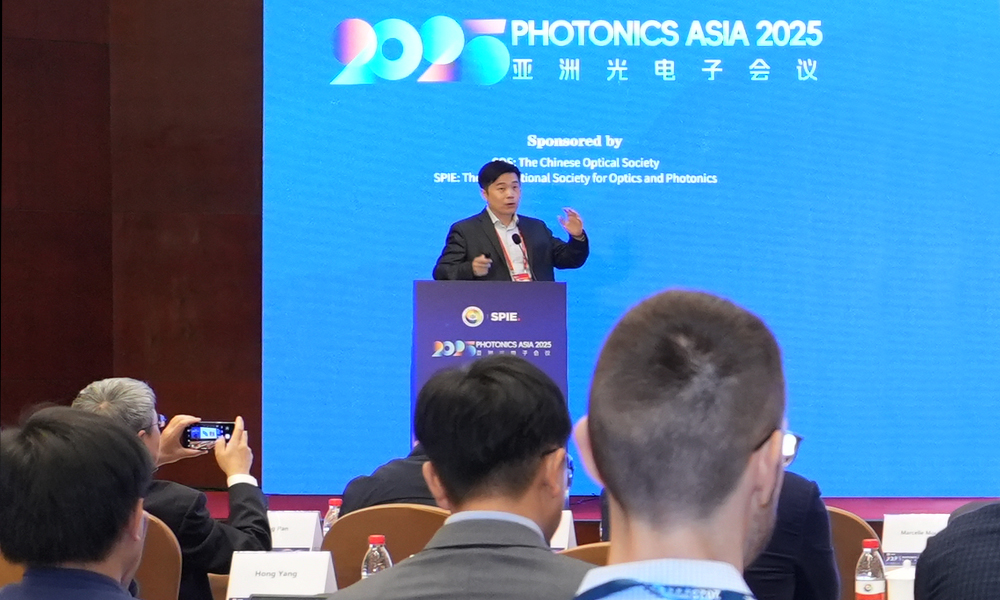 A speaker presents at a podium during Photonics Asia 2025, with the event logo and sponsor names displayed on a large blue screen behind them, while audience members listen and take photos from their seats.