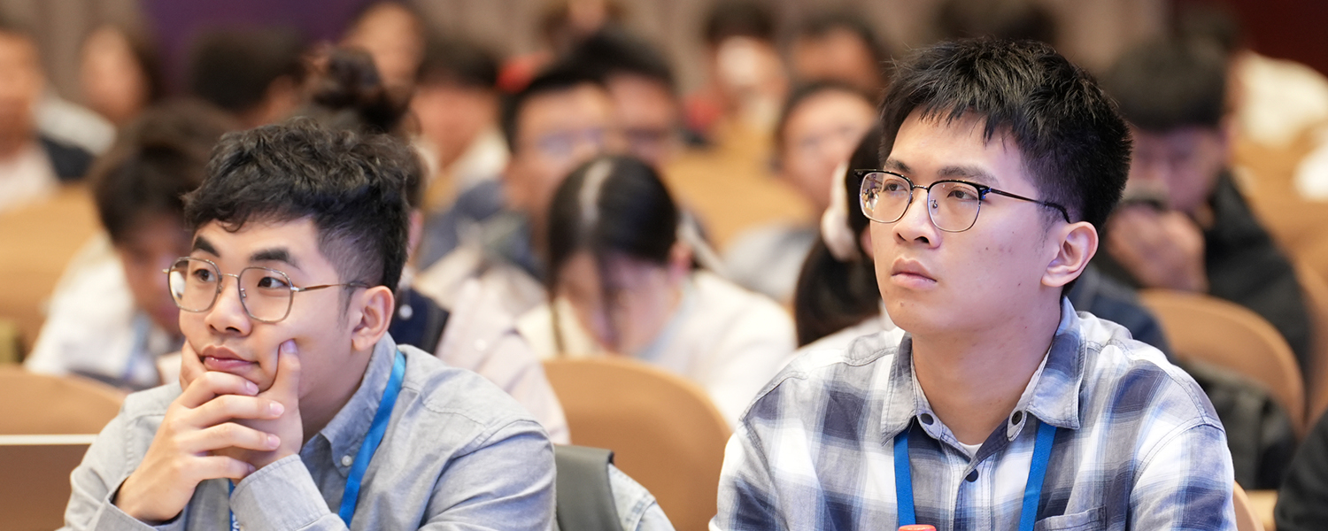 Audience members seated in a lecture hall, wearing conference badges and listening attentively to a presentation.