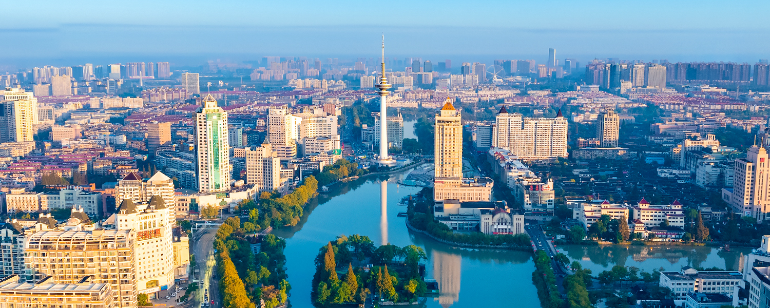 Wide aerial panorama of Nantong, China, featuring a central river lined with trees, dense urban development, modern high-rise buildings, and a landmark TV tower against a clear blue sky.