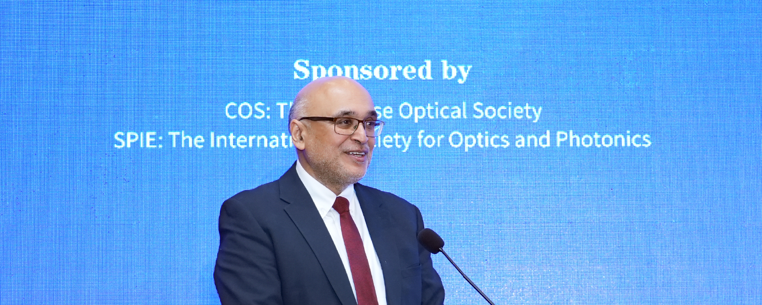 A speaker in a dark suit and red tie stands at a podium with a microphone, presenting on stage in front of a large blue screen displaying sponsor names related to optics and photonics organizations.