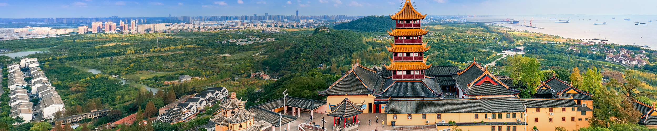 Panoramic view of Nantong, China, showing a traditional multi-tiered pagoda complex in the foreground, surrounded by green hills, with modern city buildings and the coastline visible in the distance.