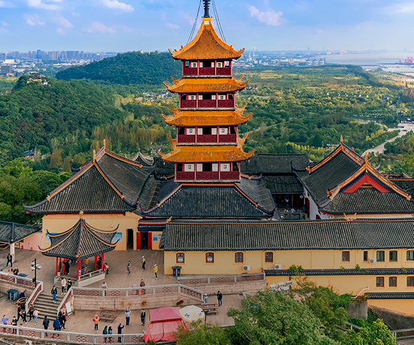 View of Nantong, China, showing a traditional multi-tiered pagoda complex in the foreground, surrounded by green hills, with modern city buildings and the coastline visible in the distance.