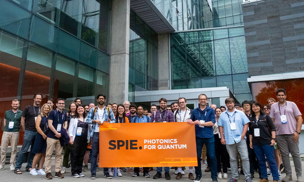 A group of conference attendees standing outdoors in front of a modern glass building, holding an orange banner that reads ‘SPIE: Photonics for Quantum.’ Participants are wearing conference badges and casual professional attire.