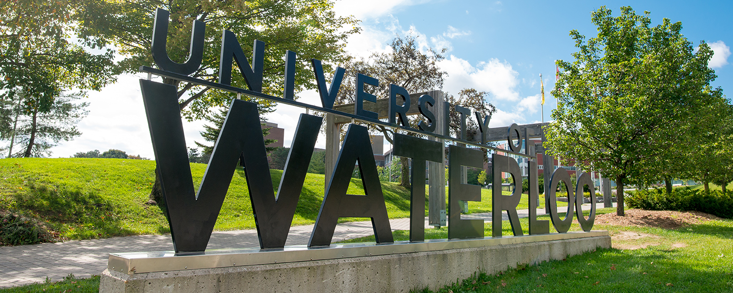 The University of Waterloo name displayed in large black metal letters along a landscaped campus walkway with trees and buildings behind it.