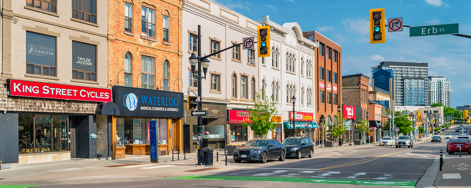 Sunny daytime view of a city street lined with historic brick and stone storefronts, including shops and medical offices, with parked and moving cars, traffic lights, and a bike lane in the foreground.