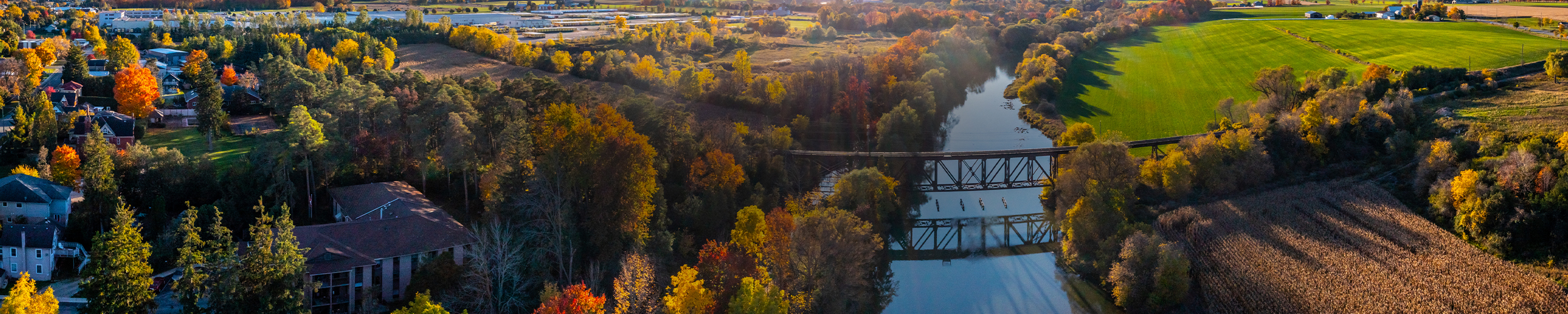 Panoramic aerial view of a river running through a forest of autumn foliage, crossed by a steel truss bridge, with residential buildings on one side and open farmland on the other.