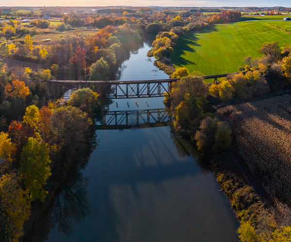 Aerial view of a river running through a forest of autumn foliage, crossed by a steel truss bridge, with residential buildings on one side and open farmland on the other.