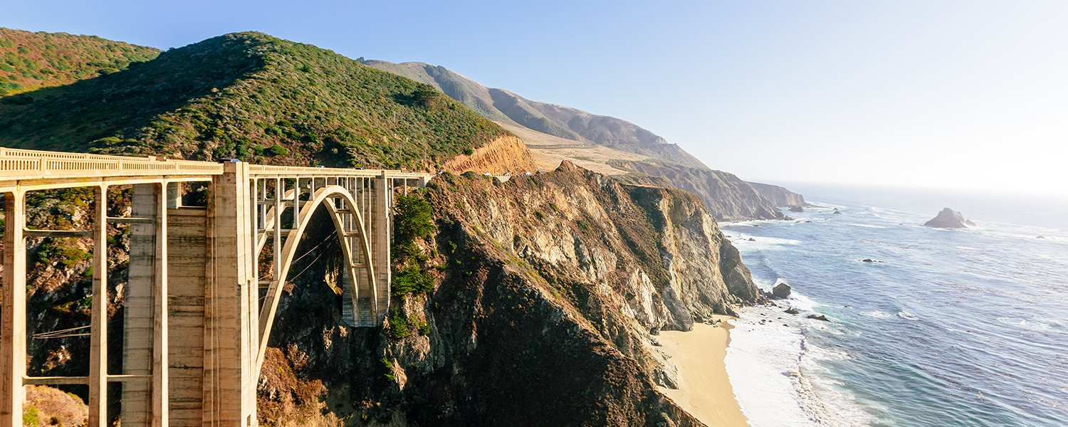 A concrete arch bridge spans a rugged coastal canyon above the Pacific Ocean, with steep green hills and crashing waves along the shoreline.