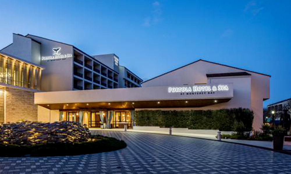 Exterior of the Portola Hotel & Spa at Monterey Bay at dusk, showing the illuminated entrance canopy, modern hotel façade, and patterned driveway under a blue evening sky.