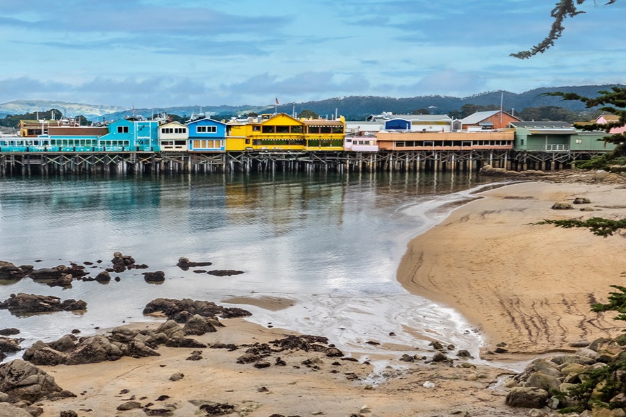 A sandy shoreline with scattered rocks curves toward calm water, backed by a row of colorful buildings on a wooden pier and low hills under a cloudy sky.