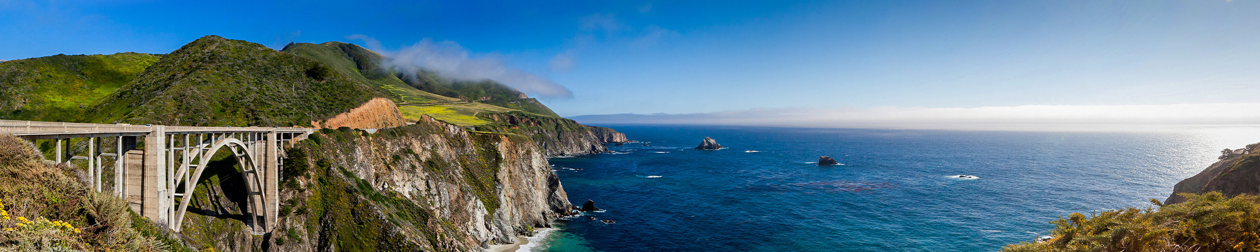 Panoramic view of Bixby Creek Bridge along the rugged Big Sur coastline near Monterey, California, with steep cliffs, green hills, and the deep blue Pacific Ocean under a clear sky.