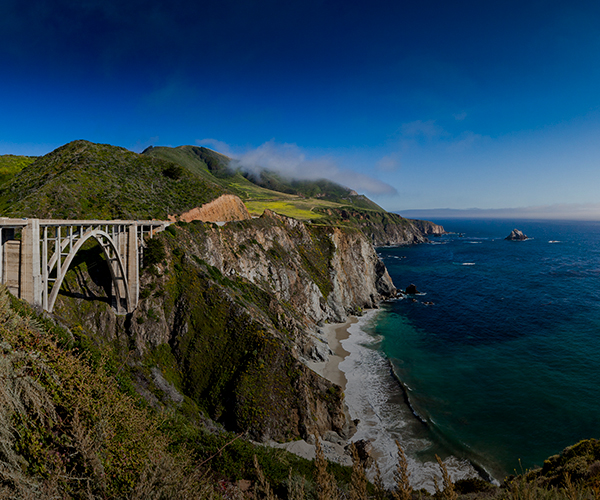 Scenic view of Bixby Creek Bridge along the rugged Big Sur coastline near Monterey, California, with steep cliffs, green hills, and the deep blue Pacific Ocean under a clear sky.