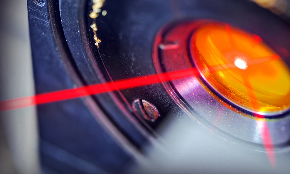 A close-up view of a red laser beam passing over an orange-tinted optical lens, with surrounding metal components slightly out of focus.