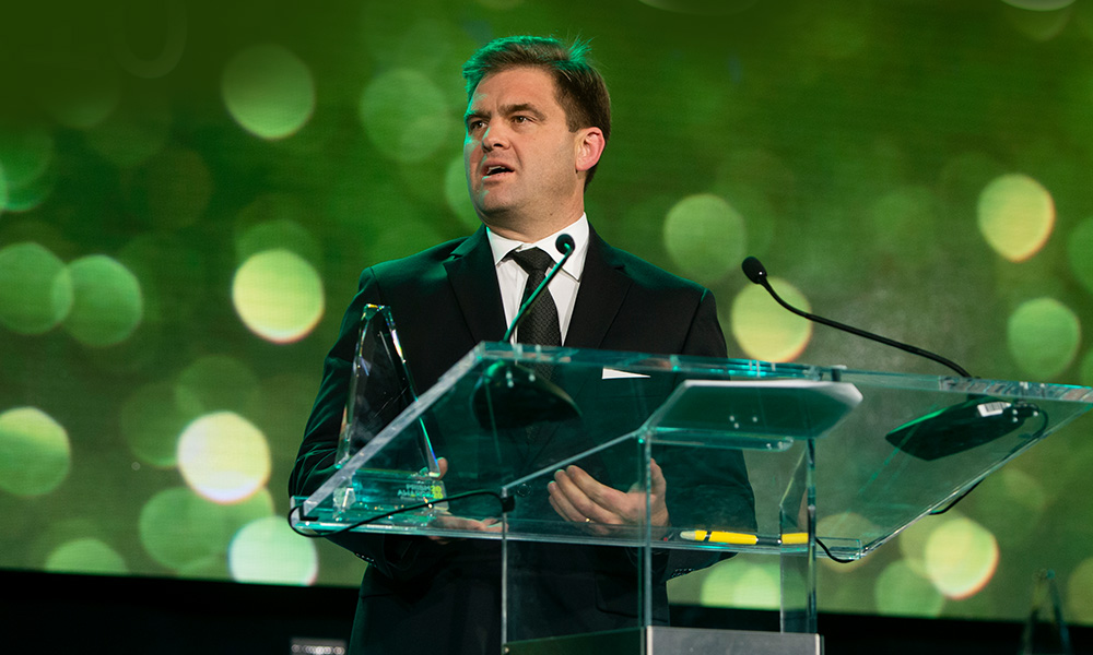 Person in a dark suit speaking at a podium with microphones, with a trophy on the podium and a green bokeh background.