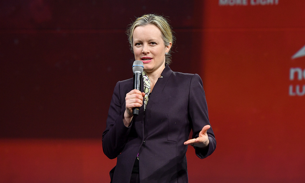 A presenter stands on a stage holding a microphone, wearing a dark blazer and light shirt, with a red background and partially visible text that says "Startup Challenge." 