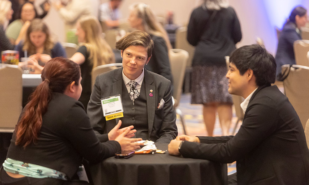 At an SPIE Member Lounge, three attendees sit closely around a small round table engaged in discussion. All wear event badges on lanyards and business‑appropriate attire. Additional attendees sit and stand at nearby tables in the background.