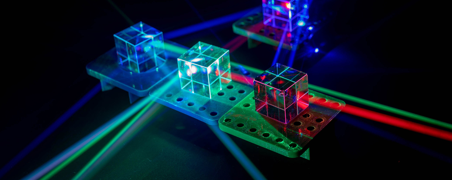 A scientific laser setup involving three transparent cubes is placed on a perforated metal platform. Each cube is illuminated by different colored laser beams, including green, blue, and red. The laser beams intersect and pass through the cubes, creating a visually striking pattern of light reflections and refractions. The background is dark, highlighting the vibrant colors of the laser beams and their interactions with the cubes.
