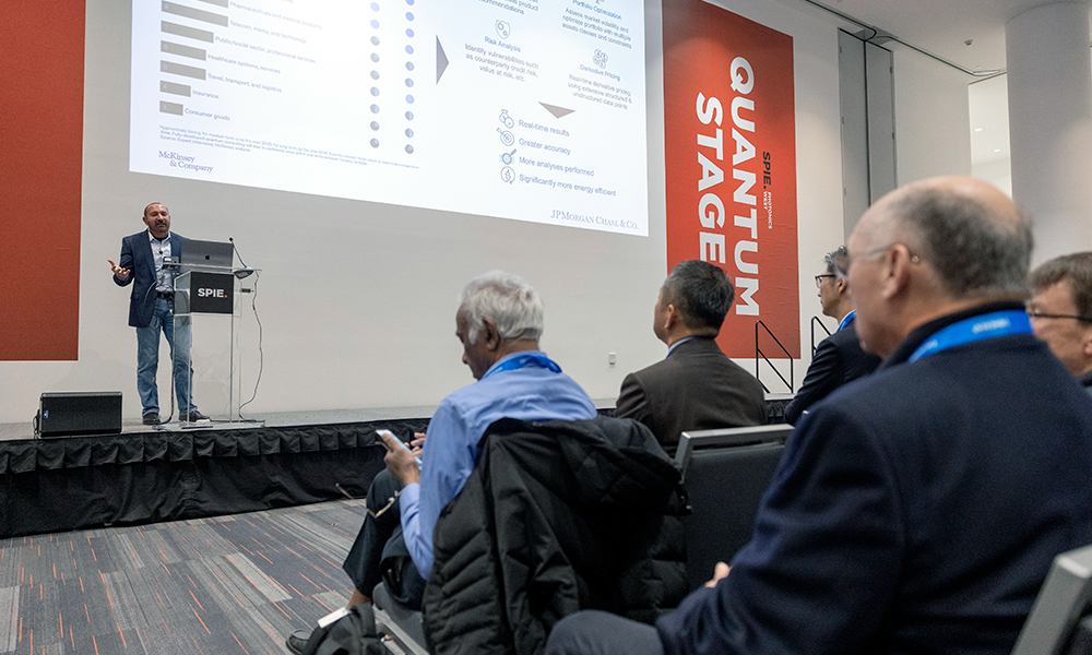 A speaker presents the Quantum West Business Summit, standing behind a podium and gesturing toward a screen filled with charts and bullet points, while attendees seated in rows listen, take notes, and use electronic devices.