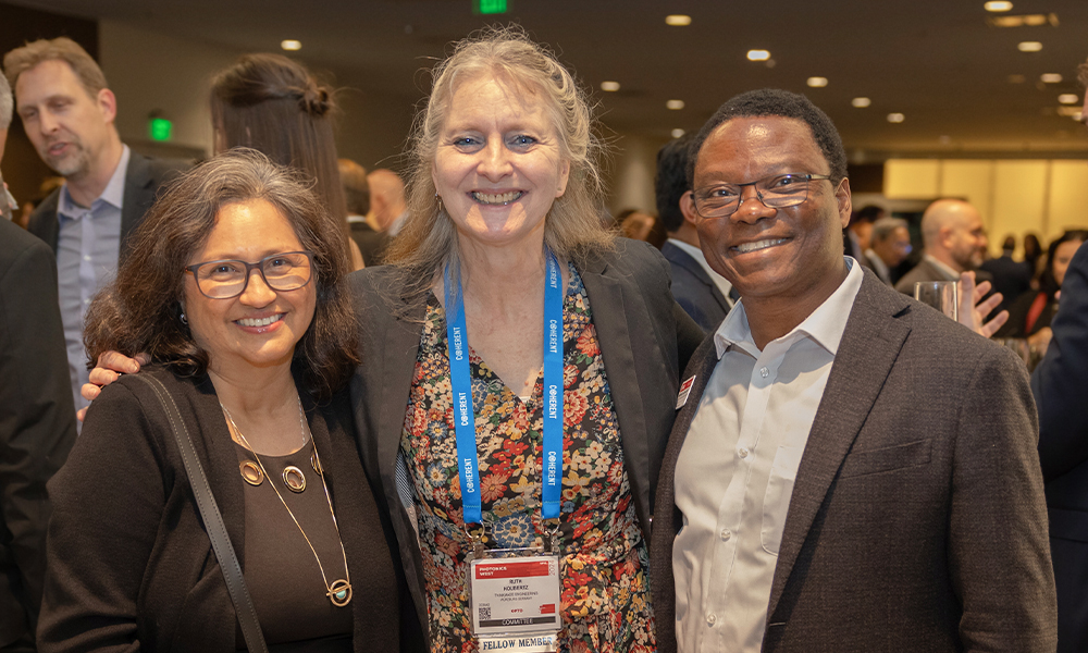 Three attendees pose together at a professional event, smiling at the camera. The central figure wears a floral dress with a black blazer and event badge, flanked by two individuals in business attire. 