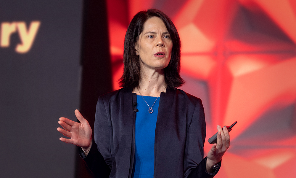Person in a blue dress and dark blazer giving a presentation holding a remote control, standing in front of a red geometric background.