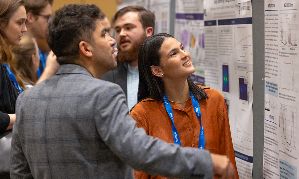 A person presents a scientific poster at a conference, pointing to a section while holding a beverage can. The poster includes labeled sections such as 'Polarimetry,' 'Results,' and 'Conclusion,' with diagrams, graphs, equations, and images illustrating the research. Another attendee listens, and others are visible in the background.
