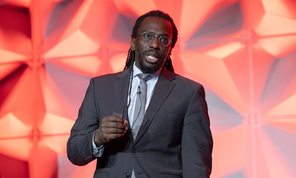 A person in a dark suit, white shirt, and tie speaks on the SPIE BiOS stage, holding a small device. The backdrop features a geometric pattern illuminated with red and pink lighting.