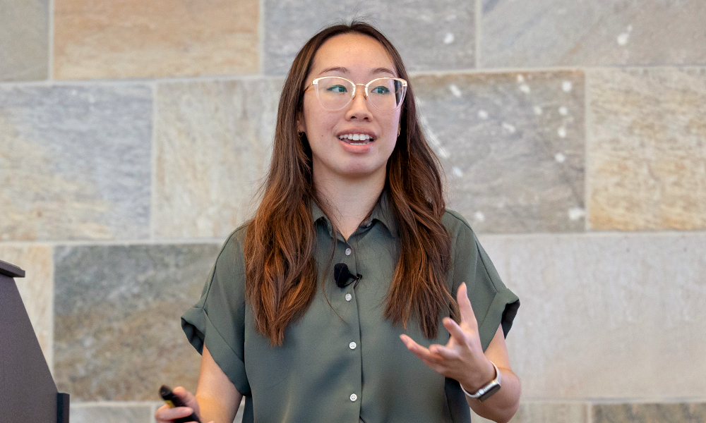 A female presenter speaks at a podium, wearing glasses, a green shirt, in front of a muted green and blue background.